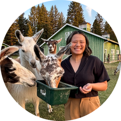 Tutor Sophia at a farm, smiling big and holding a grain bucket from which three alpacas are eating.